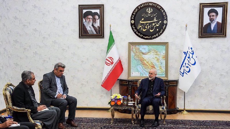 Three men in suits sit in an official room with framed portraits, an emblem reading "Islamic Parliament of Iran," an Iranian flag, and a map of the region.