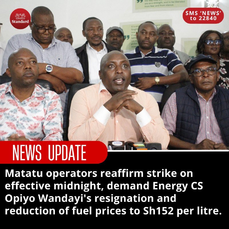 A group of men, with one in a peach shirt speaking at microphones, sit together during what appears to be a press briefing, with The Standard newspaper branding visible.