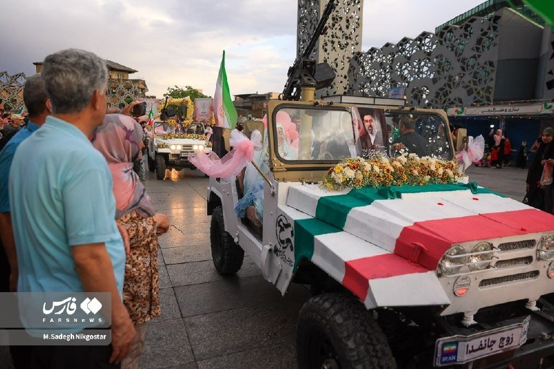 A decorated white vehicle draped in a green, white, and red flag leads a procession down a public square, with a portrait displayed in the windshield and onlookers watching.