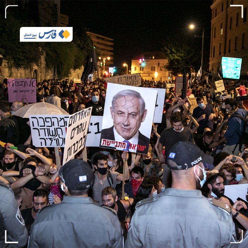 A nighttime street protest shows a large crowd holding signs with Hebrew text and a cracked-face portrait, facing police officers in caps labeled "POLICE."