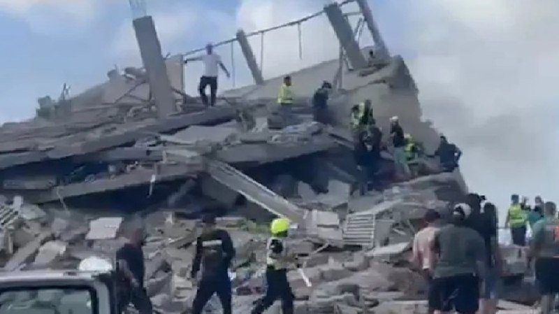 Rescue workers in high-visibility vests and bystanders gather atop and around a collapsed concrete building amid a large debris pile.