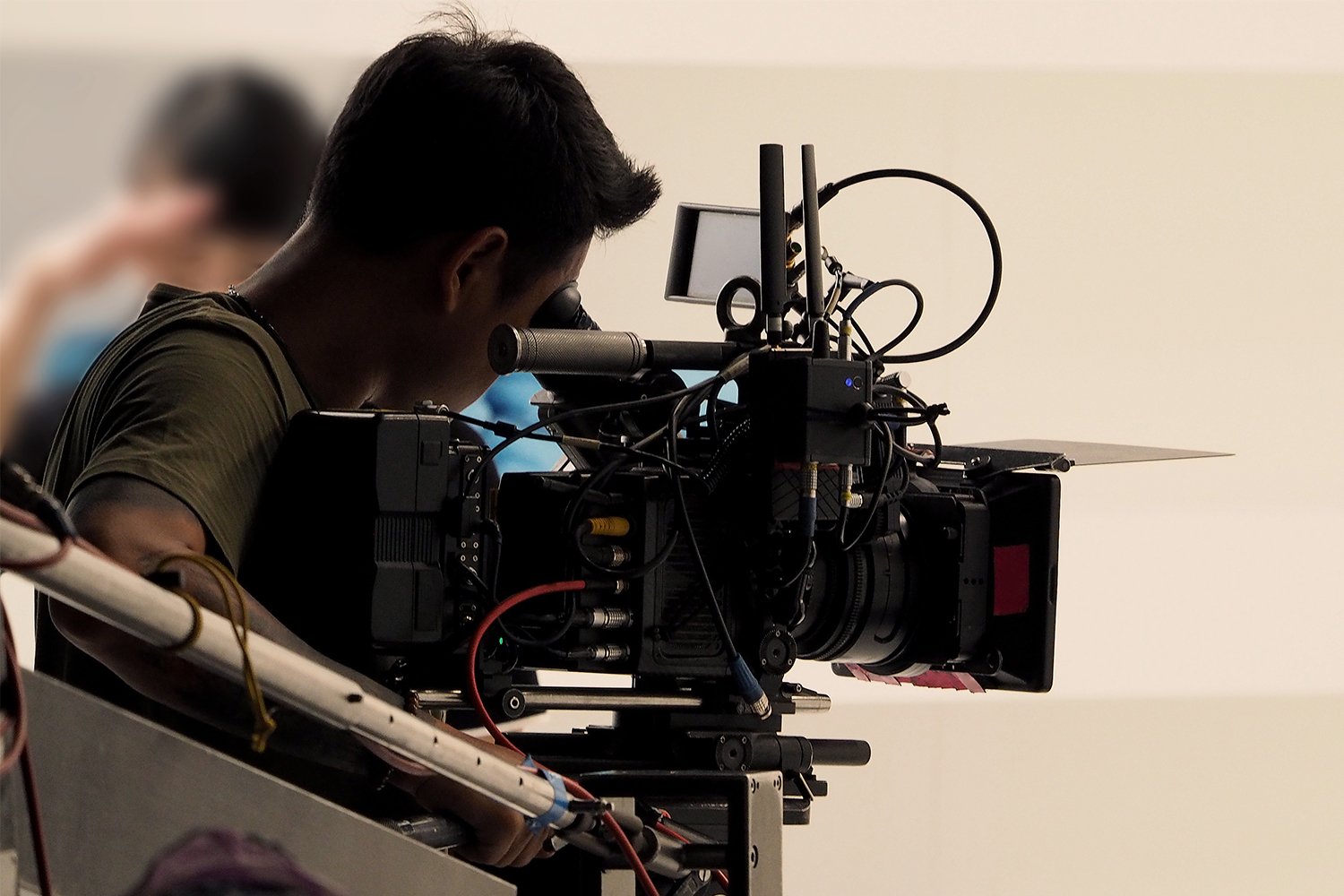 A man in an olive green shirt looks through the viewfinder of a large professional video camera equipped with a wireless transmitter, monitor, and matte box.