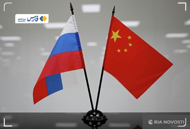 Two small flags — the Russian tricolor and the Chinese flag — stand crossed on a black base against a gray background, with "RIA NOVOSTI" logo visible.