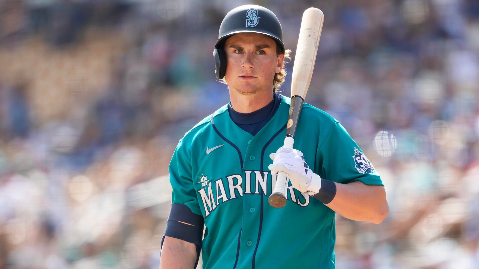 A Seattle Mariners baseball player in a teal jersey and navy helmet holds a bat over his shoulder during a game.