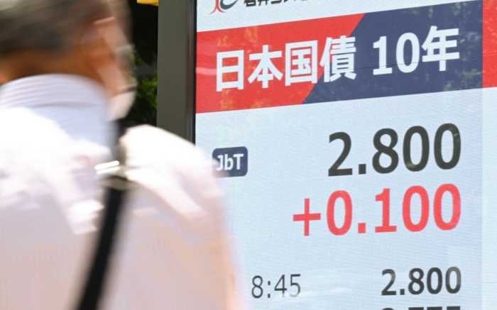 A woman wearing glasses stands near a digital display showing Japanese text reading "日本国債 10年" with the figures 2.800 and +0.100.