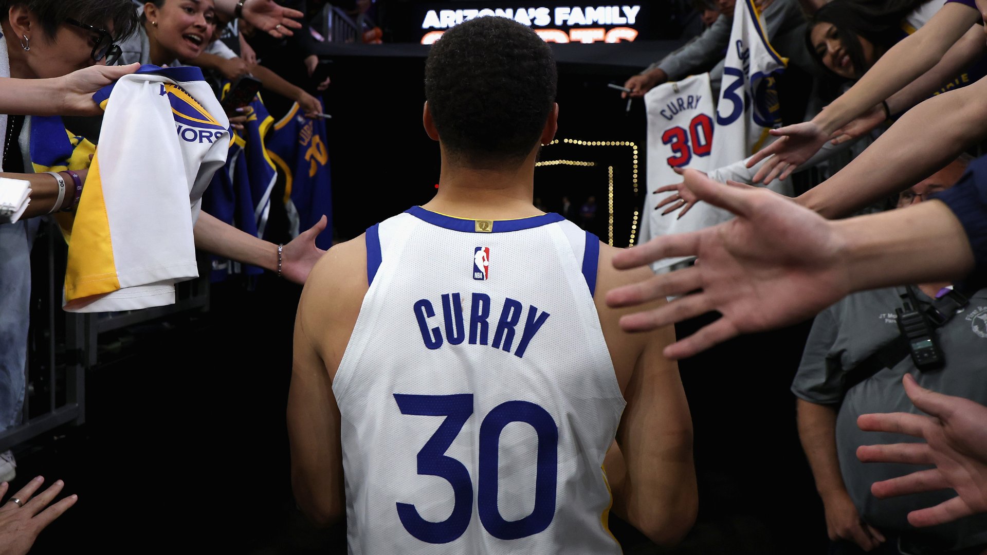 A basketball player wearing a white "Curry 30" jersey walks through a tunnel toward a court, reaching out to fans holding jerseys on either side.
