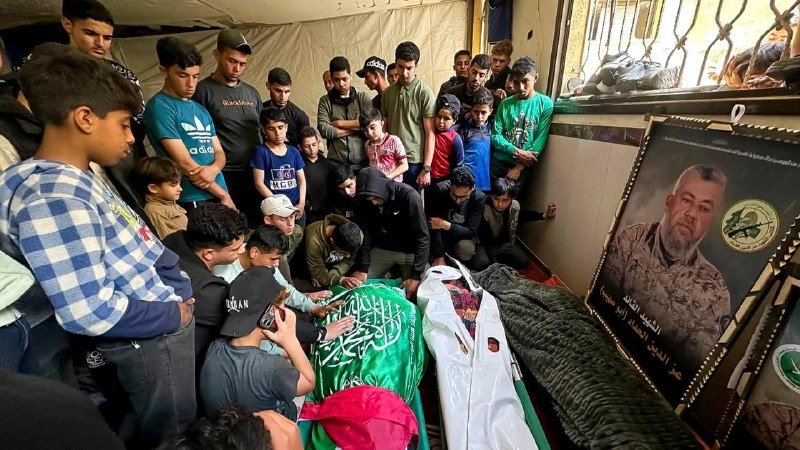 Mourners, mostly young men and boys, gather around a body draped in green and white flags inside a tented space beside a framed portrait of a man.