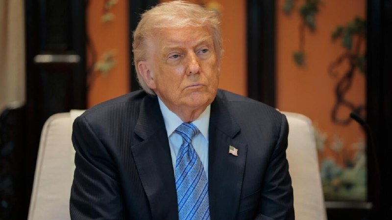 A blonde-haired man in a dark suit and blue patterned tie sits with a serious expression against an orange-toned ornate backdrop.