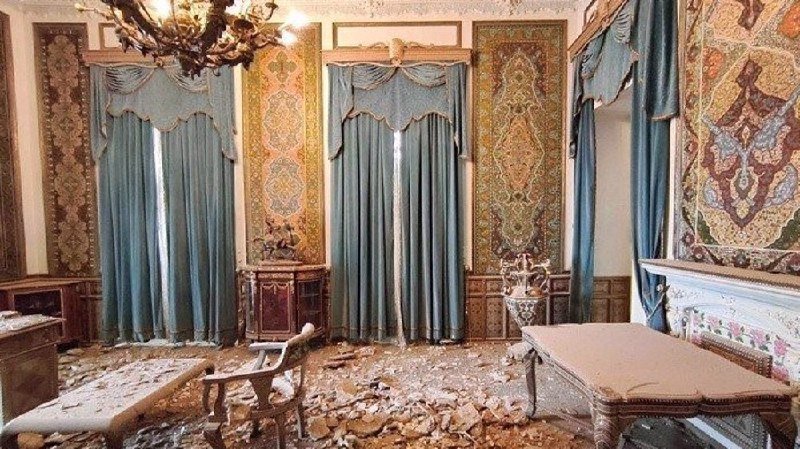 An ornate room with blue curtains, a chandelier, and antique furniture is covered in debris and broken plaster, indicating damage.