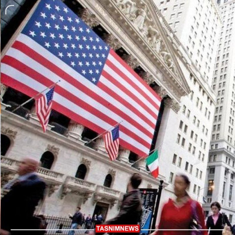 A large American flag drapes over a neoclassical building facade alongside smaller U.S. and Italian flags, as pedestrians walk on the street below.
