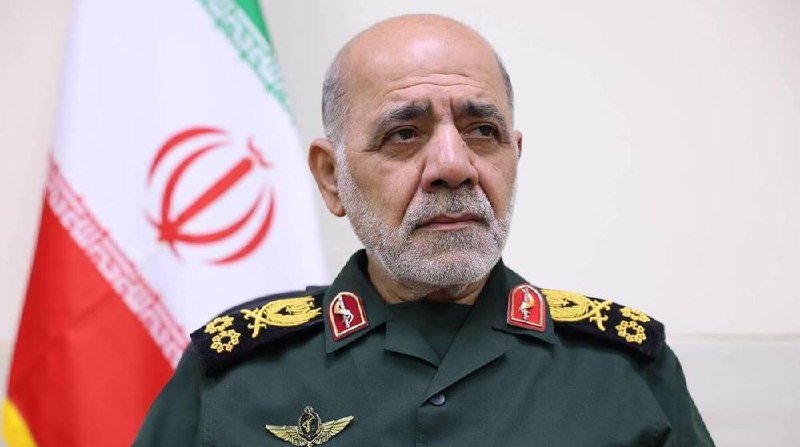 A bald, gray-bearded man in a green military uniform with gold insignia stands before a flag displaying red, white, and green colors with a central emblem.