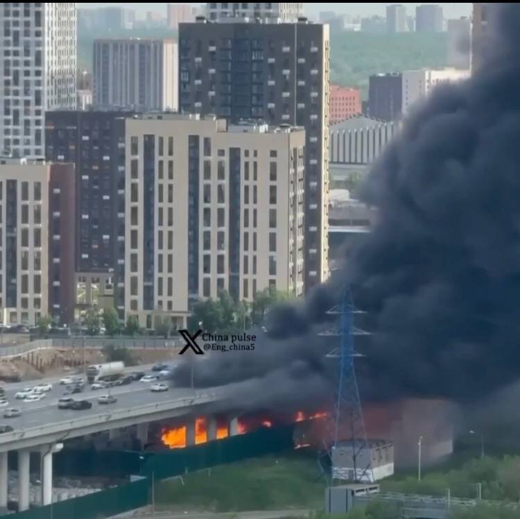 Thick black smoke billows from a fire burning beneath an elevated highway in a city with high-rise apartment buildings.