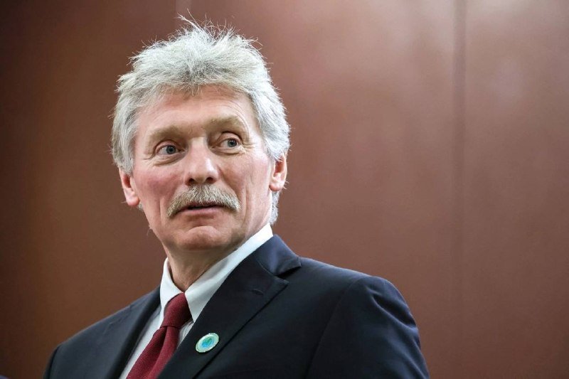 A gray-haired man with a mustache wearing a dark suit, white shirt, red tie, and a circular lapel pin stands against a brown background.