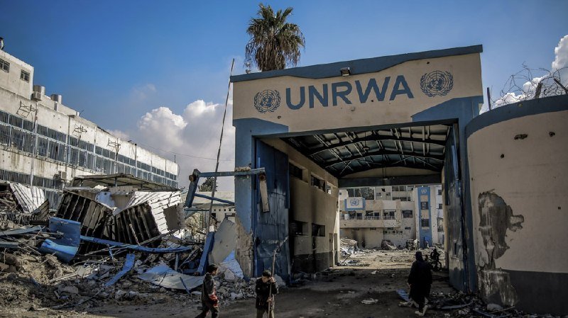 People walk through a heavily damaged compound featuring a blue and white gate marked "UNRWA" amid scattered debris and partially destroyed buildings.