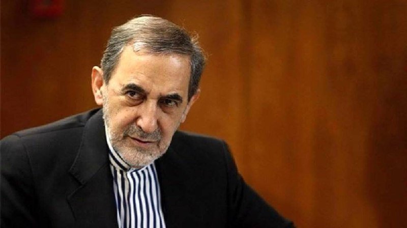 A gray-haired, bearded man wearing a dark suit jacket and striped shirt looks toward the camera against a wooden background.