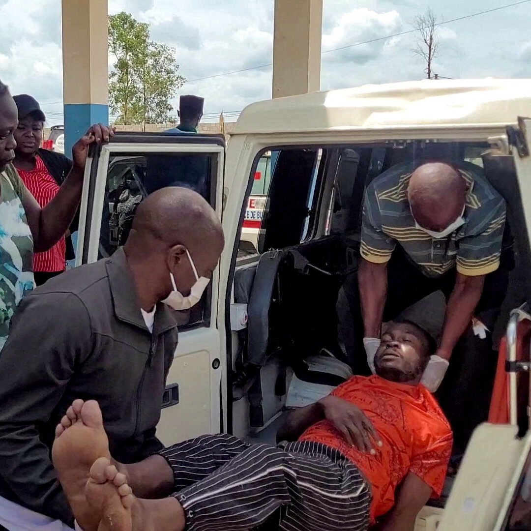 Several men load an unconscious person in an orange shirt and striped trousers into the back of a white van, with onlookers gathered nearby.