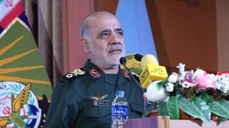 A gray-bearded man in a dark green military uniform with insignia speaks at a podium with microphones, water bottle, and floral arrangement, with an emblem and Iranian flag visible behind him.
