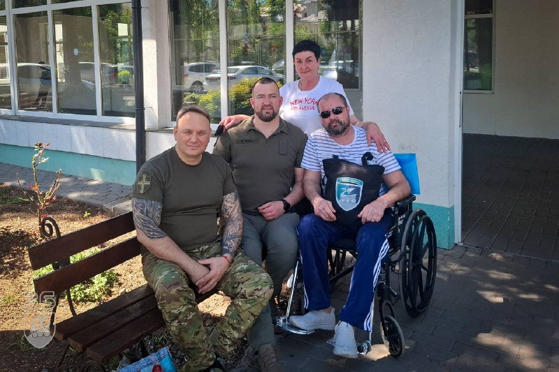 Four people pose together outside a building with large windows — two men sit on a wooden bench, one man in a wheelchair wearing a "73" pouch, and a woman stands behind them.