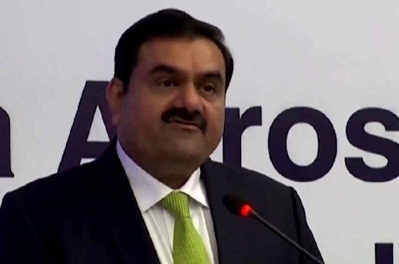 A man with a mustache in a dark suit and green tie speaks at a microphone in front of a backdrop displaying partial "Adani" lettering.