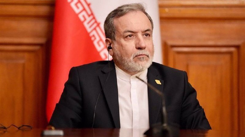A gray-bearded man in a dark suit and white shirt wears an earpiece while seated at a desk with a microphone, an Iranian flag visible behind him.