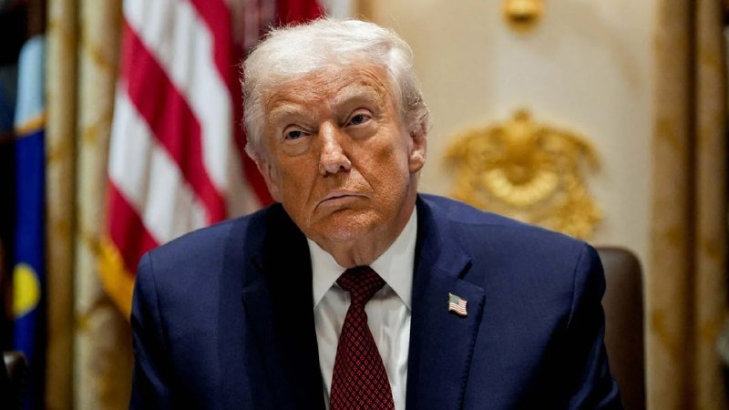 I can't identify the person in this image. I can describe that an older man with white hair wearing a dark suit, red tie, and American flag lapel pin sits in front of U.S. flags and ornate decor.