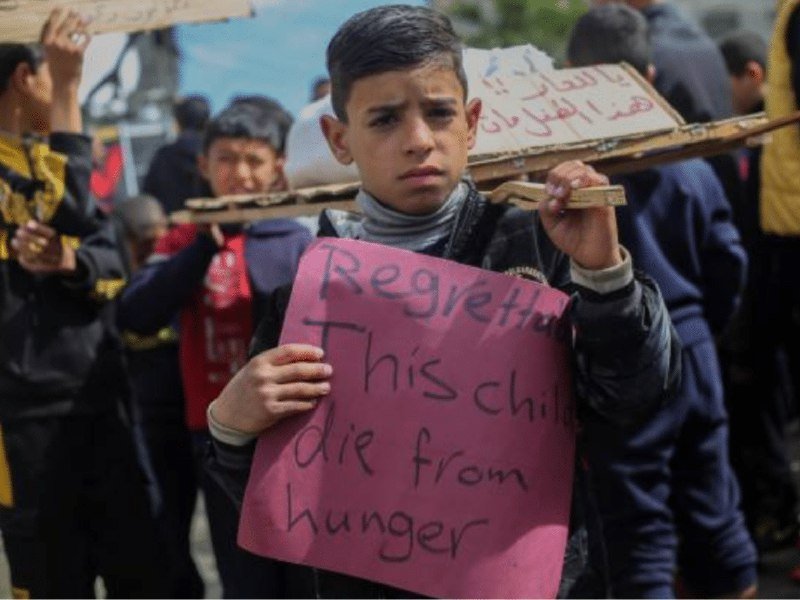 A boy holds a pink sign reading "Regretted. This child die from hunger" while standing in a crowd holding cardboard placards.