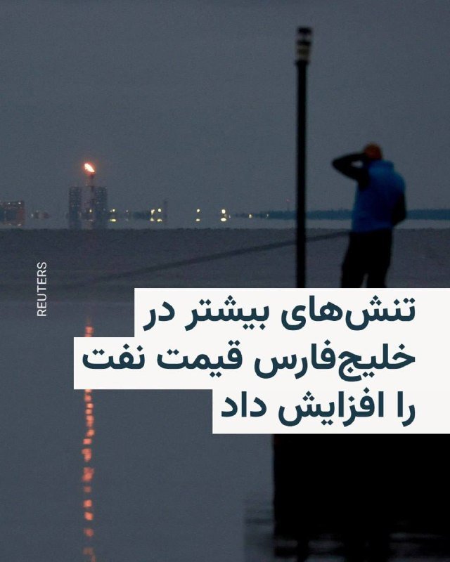 A blurred figure in a blue jacket stands on a pier at dusk, overlooking a body of water with a gas flare and lit structures visible across the shore.