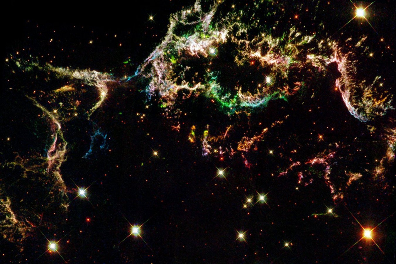 Colorful filamentary nebula structures in greens, oranges, pinks, and blues spread across a dark starfield punctuated by bright, cross-shaped stars.