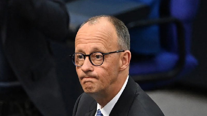 A balding man in a dark suit, white shirt, and glasses looks sideways with pursed lips at a formal indoor gathering.