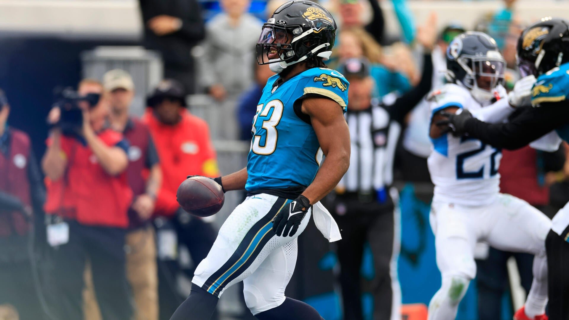 A Jacksonville Jaguars player in a teal jersey (#23) celebrates while holding a football, with a teammate and opposing players visible in the background.