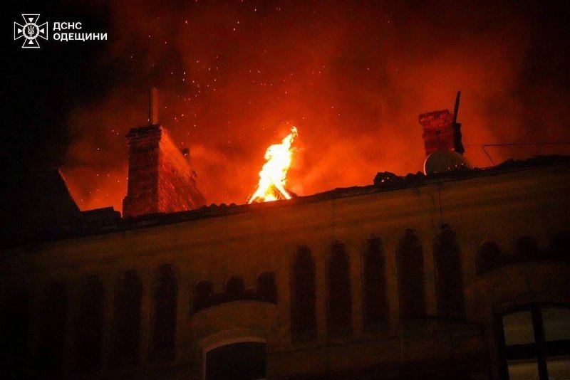 Flames and sparks erupt from the roof of a building at night, illuminating nearby chimneys and an adjacent rooftop.