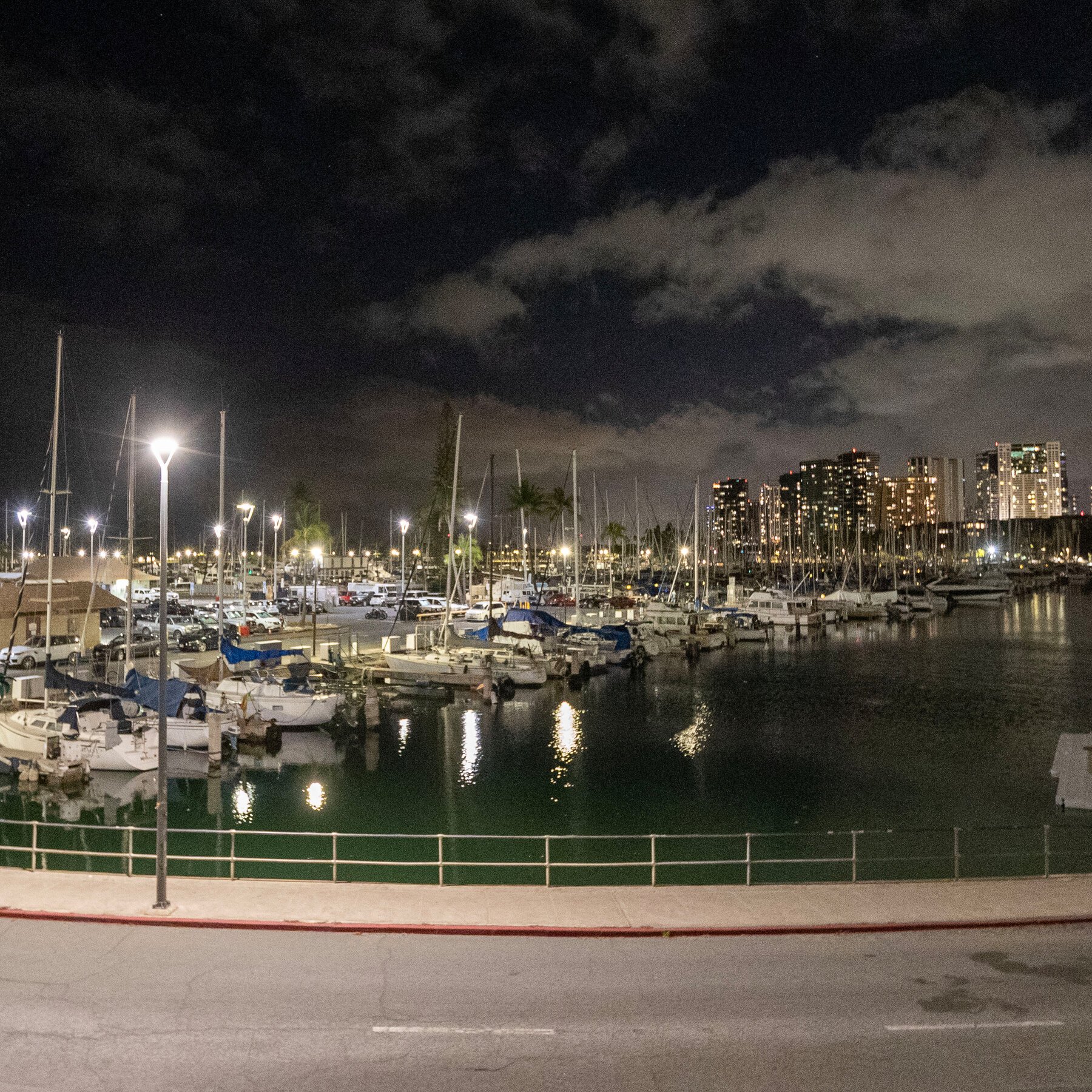 A marina filled with docked sailboats is illuminated by streetlights at night, with a city skyline of lit high-rise buildings visible in the background under a cloudy sky.
