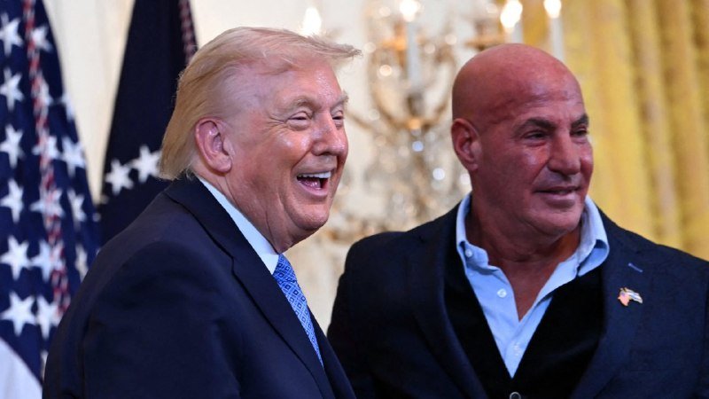 A smiling blonde-haired man in a dark suit stands beside a bald man in a dark jacket, with American flags and a chandelier visible in the background.