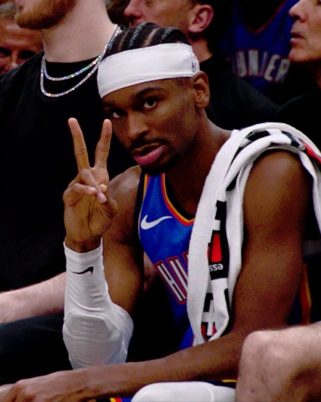 A basketball player wearing a blue jersey, white headband, and wristband flashes a peace sign toward the camera while seated on the bench.