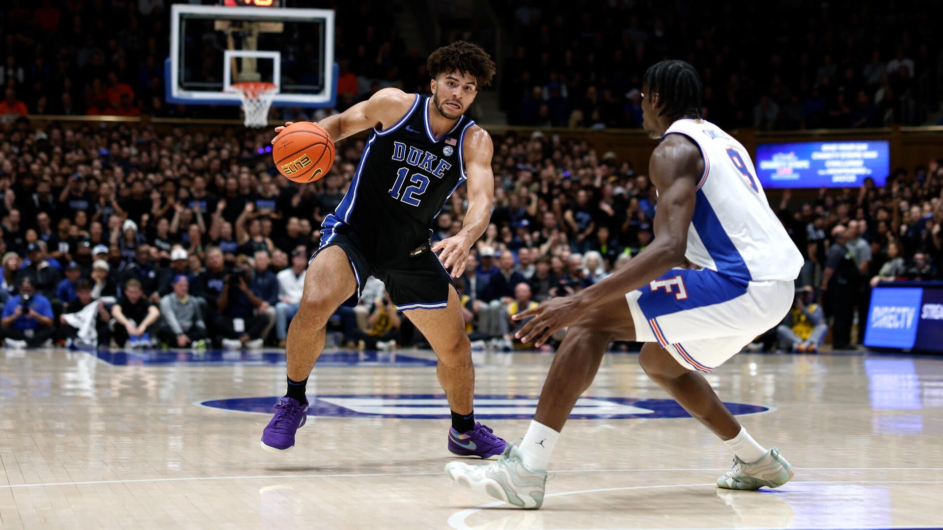 A Duke basketball player wearing a black jersey with the number 12 dribbles the ball while being defended by an opposing player in a white and blue uniform during a game.