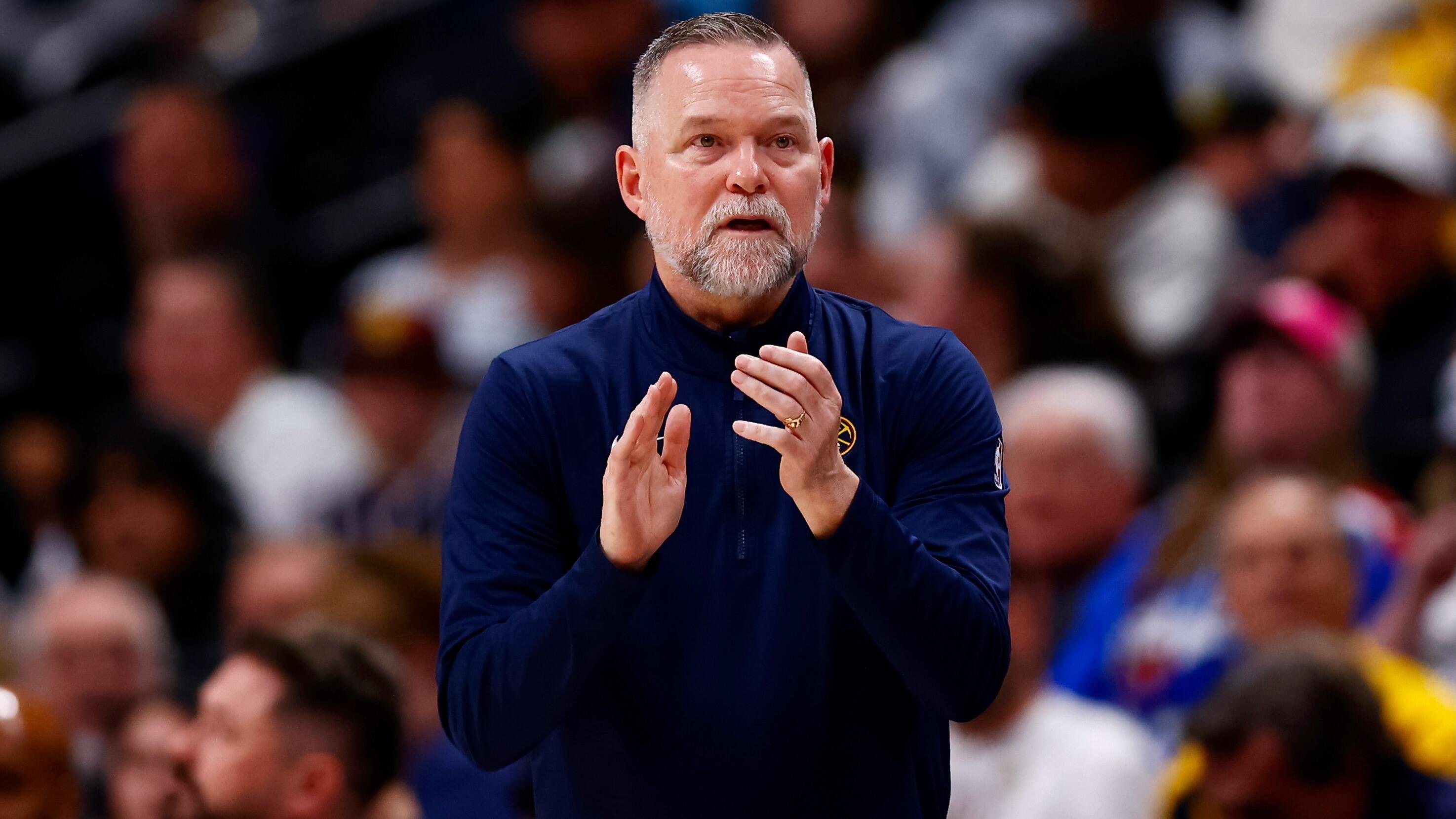 A bearded man in a navy quarter-zip jacket claps his hands while standing in front of a blurred crowd.