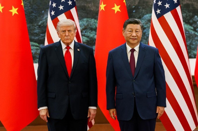 Two men in dark suits stand side-by-side facing forward, posed before alternating Chinese and American flags.