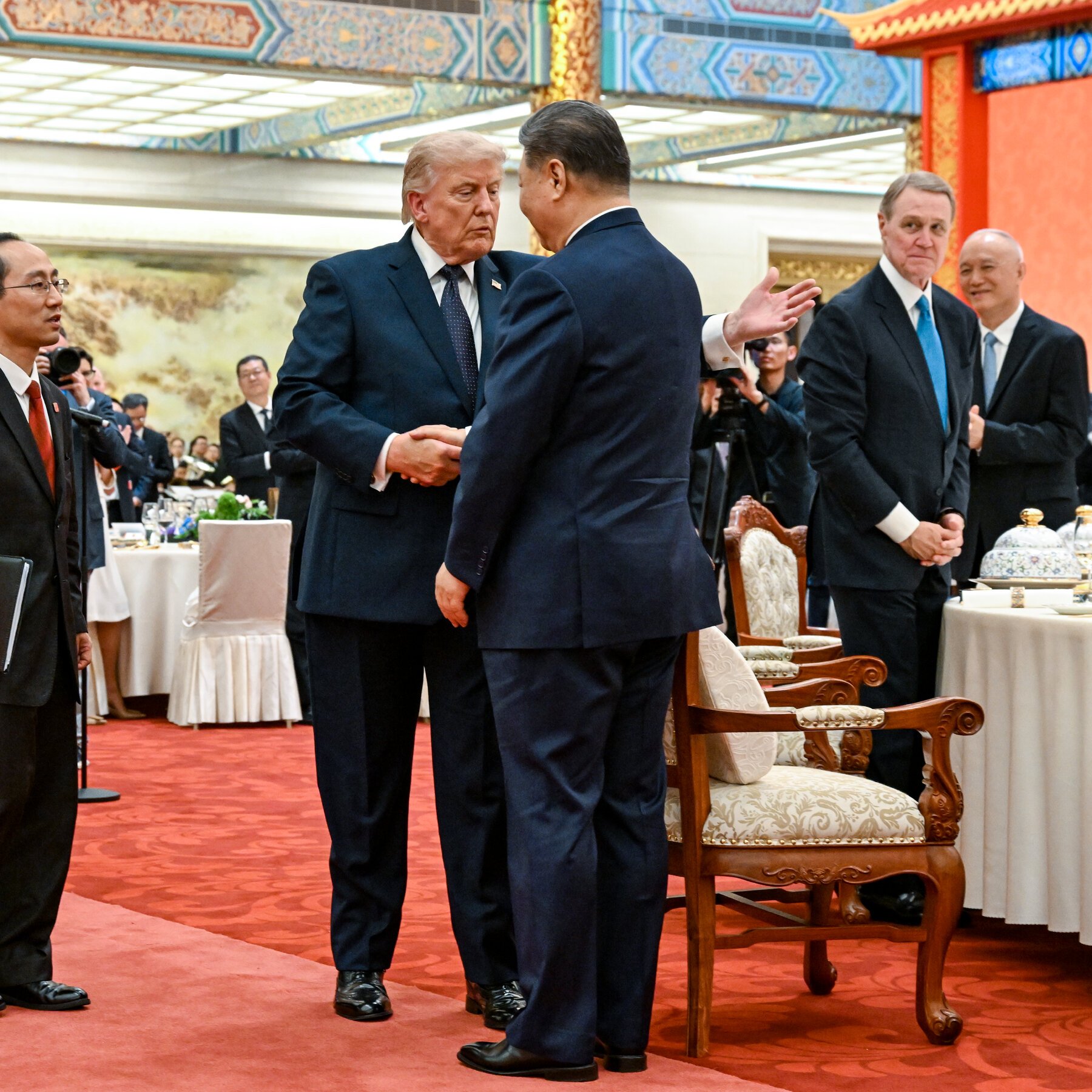 Two men in dark suits shake hands on a red carpet in an ornate hall with traditional decorative ceiling beams, while other formally dressed officials observe nearby.