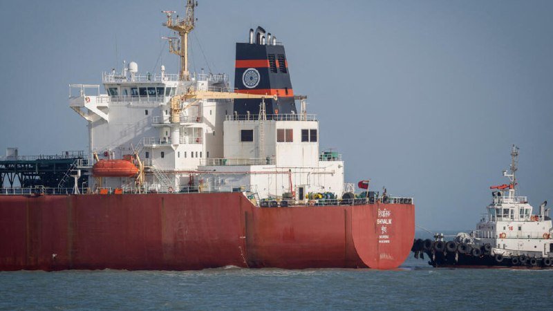 A large red-hulled cargo ship with a white superstructure and black funnel is assisted by a smaller tugboat on open water under a clear sky.