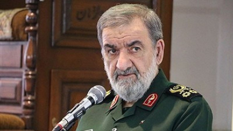 A bearded man in a green military uniform with insignia speaks into a microphone in front of a wooden paneled background.