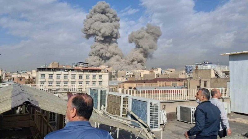 Two large plumes of gray smoke rise over a city skyline, observed by men on a rooftop with air conditioning units.