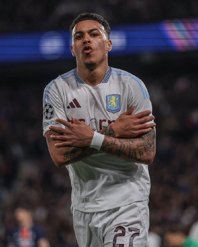 A soccer player in a white Aston Villa jersey with number 22 crosses his tattooed arms in a goal celebration during a match.
