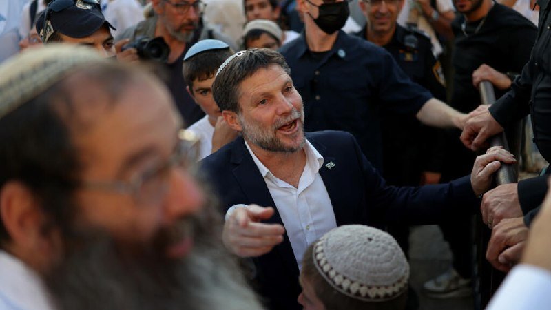 A man in a suit and white shirt, wearing a small skullcap, speaks amid a crowd of people, some wearing kippot, with photographers and others gathered around.