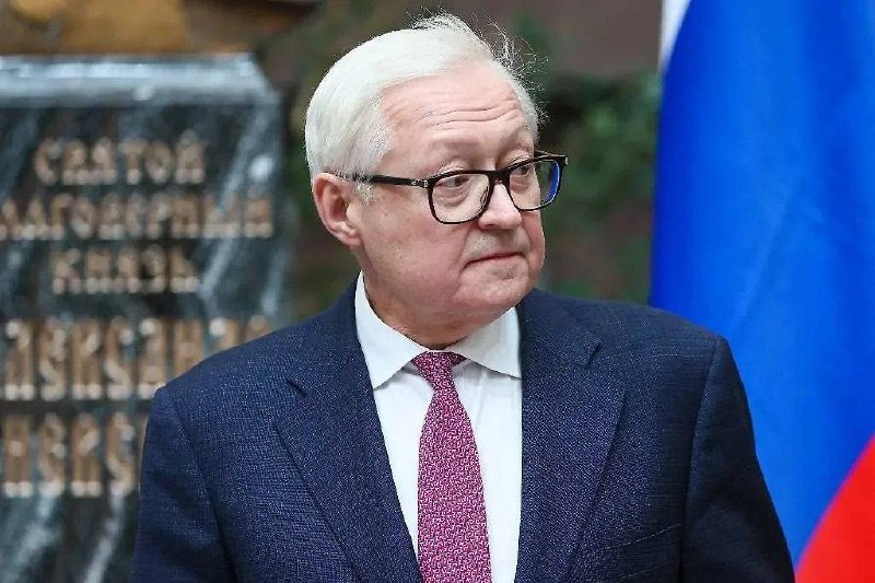 An older man with white hair and glasses, wearing a dark suit and pink tie, stands near a Russian flag with a partially visible engraved monument behind him.