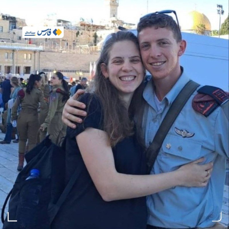 A smiling woman in a black dress embraces a man in a light blue military uniform near a stone plaza, with a golden-domed building and other uniformed personnel visible in the background.