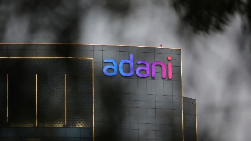 A modern building facade displays an illuminated "adani" sign in blue and pink lettering against a cloudy sky.