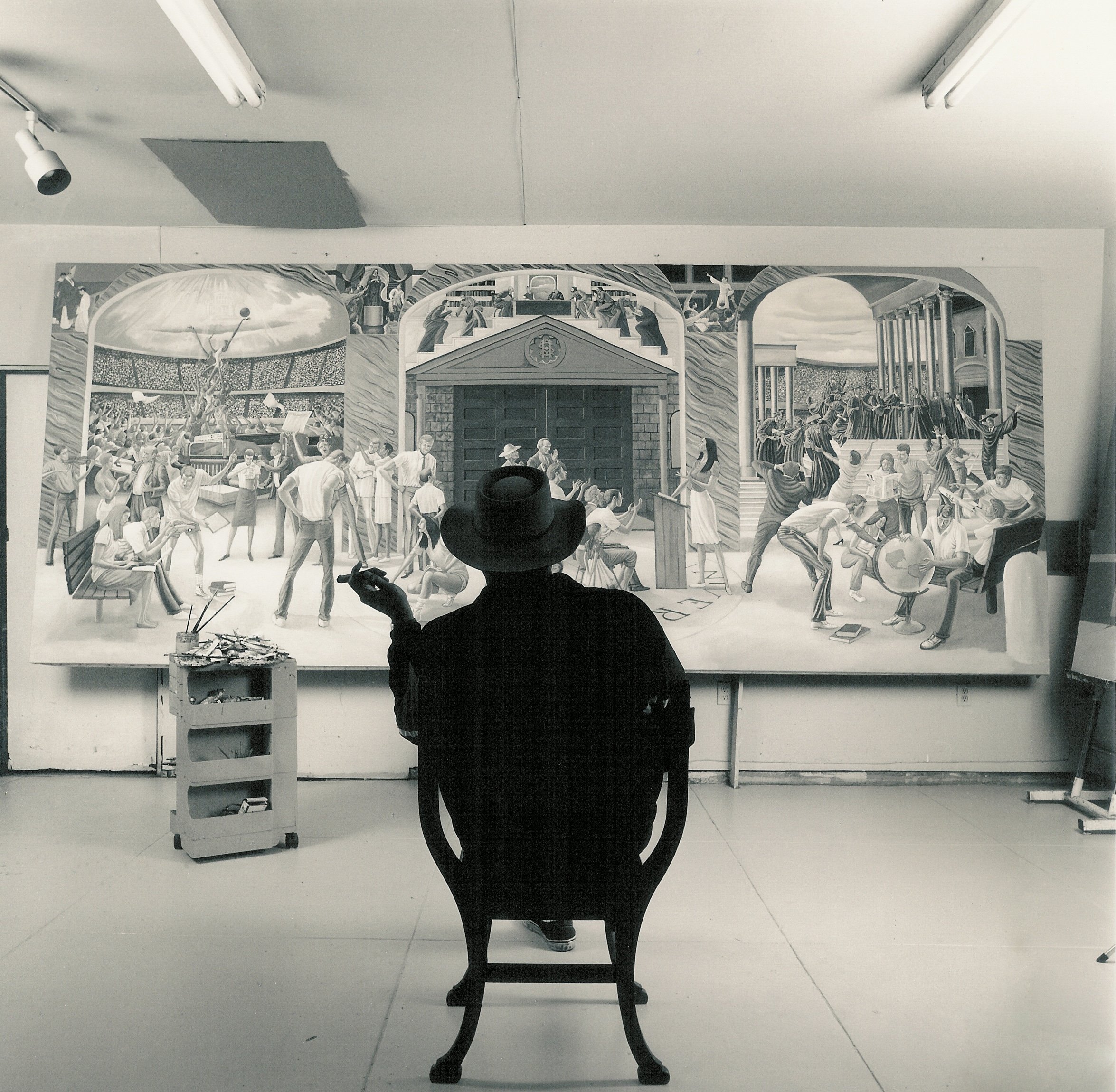 A silhouetted figure in a wide-brimmed hat sits in a chair, facing a large black-and-white mural depicting crowds, a domed building, and classical columns in a brightly lit room.