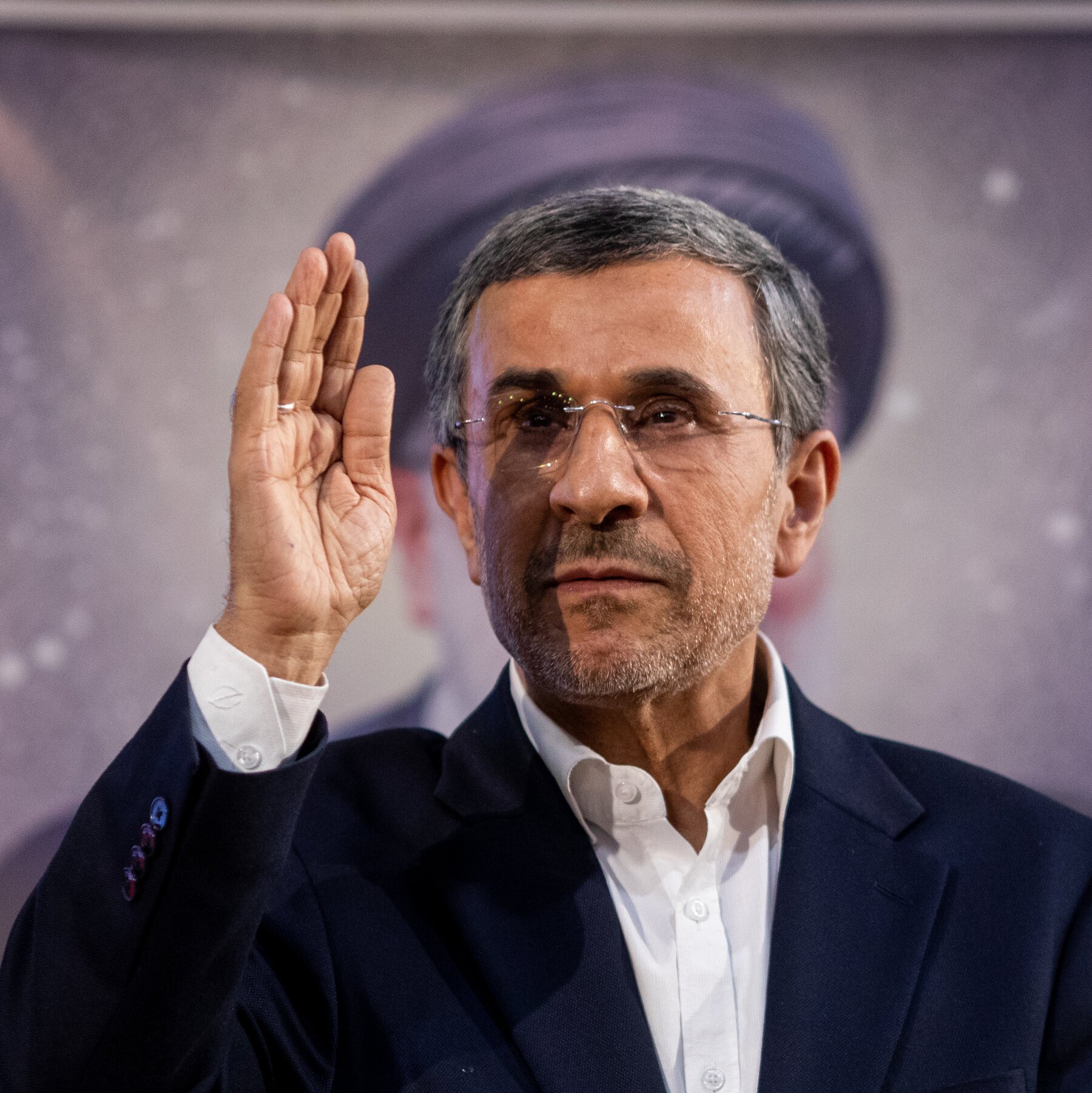 A gray-haired man in glasses and a dark suit raises his right hand in a waving gesture against a blurred purple-toned backdrop.