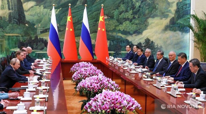 Two delegations in dark suits sit facing each other at a long conference table adorned with purple orchids, with Russian and Chinese flags displayed between them.