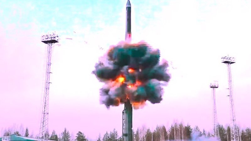 A missile launches skyward amid a large fireball and smoke, flanked by tall light towers against a pale pink sky.